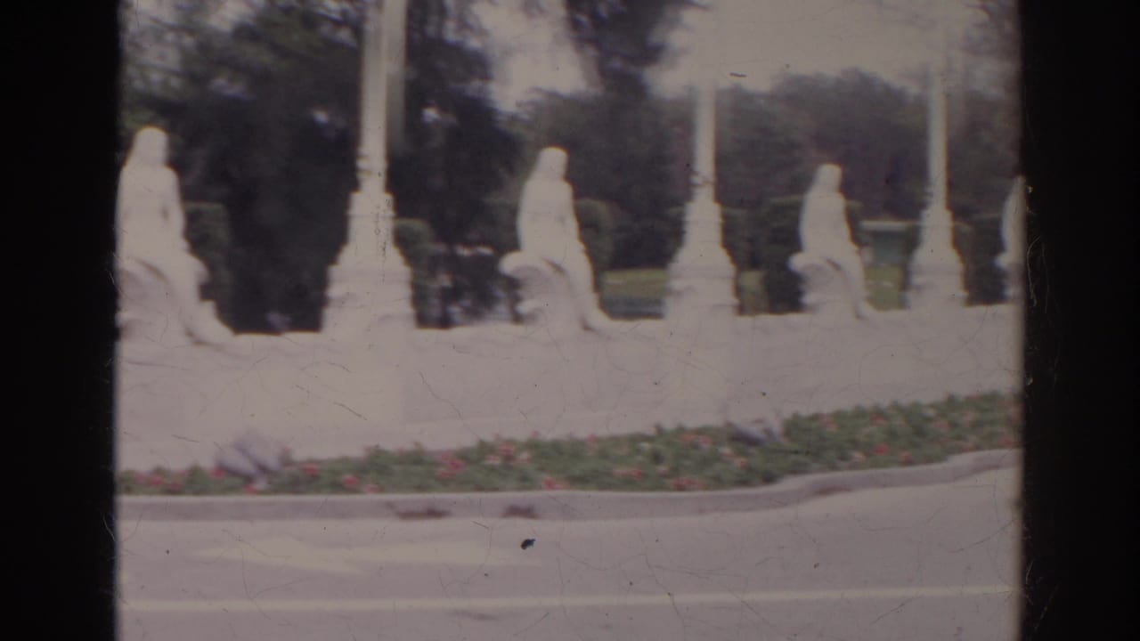 1966 Norfolk frame with white fountain figures, tall poles, clipped hedges, flowers, and a roadway in the foreground.