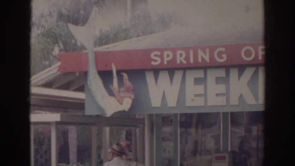 1966 home-movie frame with partial attraction or storefront signage, flags, and a pedestrian in the lower foreground.