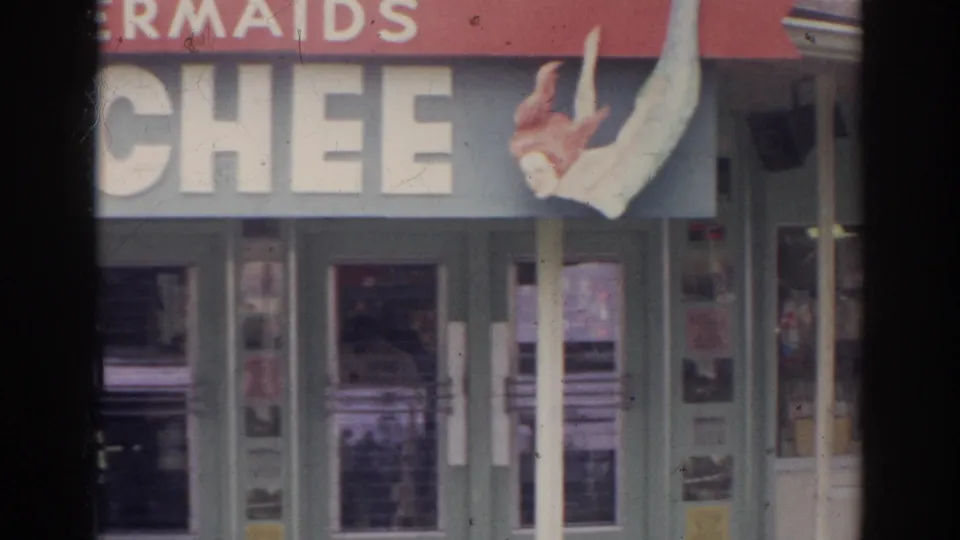 1966 Norfolk home-movie frame showing a partial storefront sign with a mermaid figure above glass entrance doors.