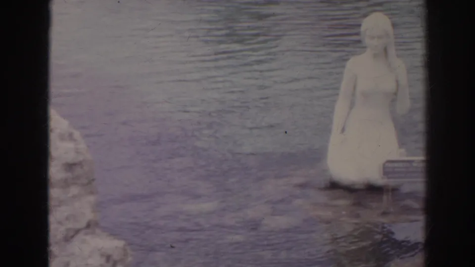 1966 Norfolk frame showing a pale female figure statue in the water with a small sign at its base and rocky shoreline at left.