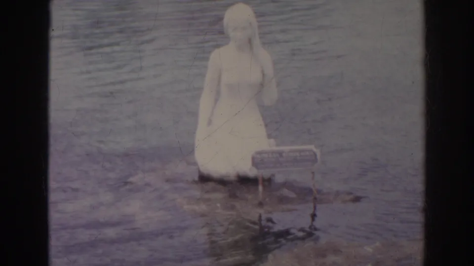 1966 Norfolk 8mm frame showing a woman in white standing in shallow lake water near a small sign.