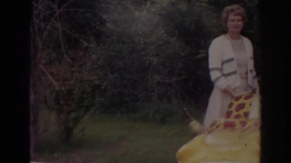 1966 Norfolk home-movie frame with a woman in a cardigan near brush and a large yellow red-spotted inflatable creature.
