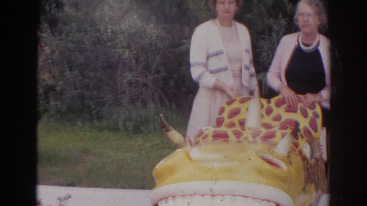 1966 Norfolk frame showing two women in dressy clothing behind a yellow red-spotted inflatable creature near lakeside brush.
