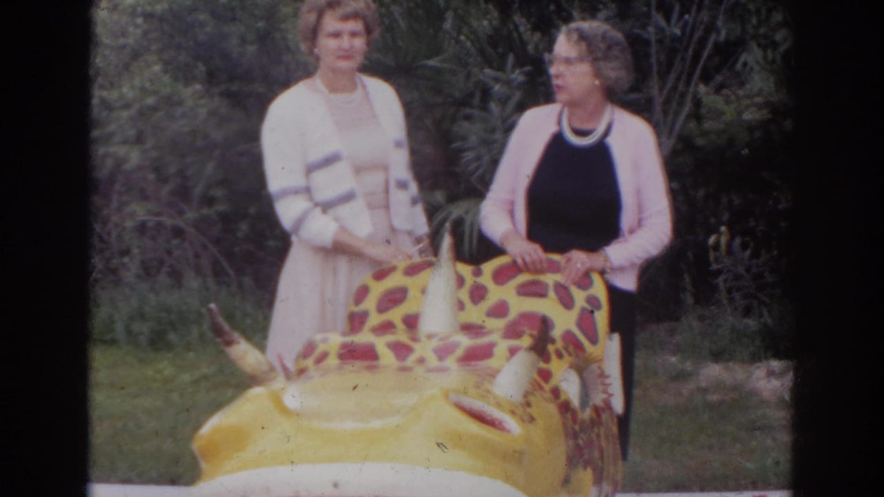 Two women stand beside a yellow spotted dinosaur ride in a grassy area.