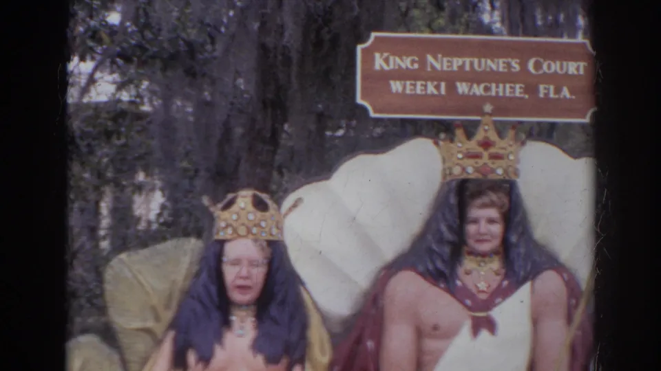 Two adults pose below a King Neptune's Court Weeki Wachee sign, framed by shell-like props and trees in daylight.