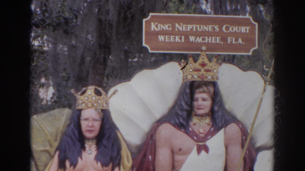 Two crowned figures pose beneath a King Neptune's Court sign in front of a shell backdrop.
