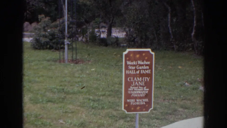 A Weeki Wachee Star Garden Hall of Fame sign honoring Calamity Jane stands on grass with trees behind it.