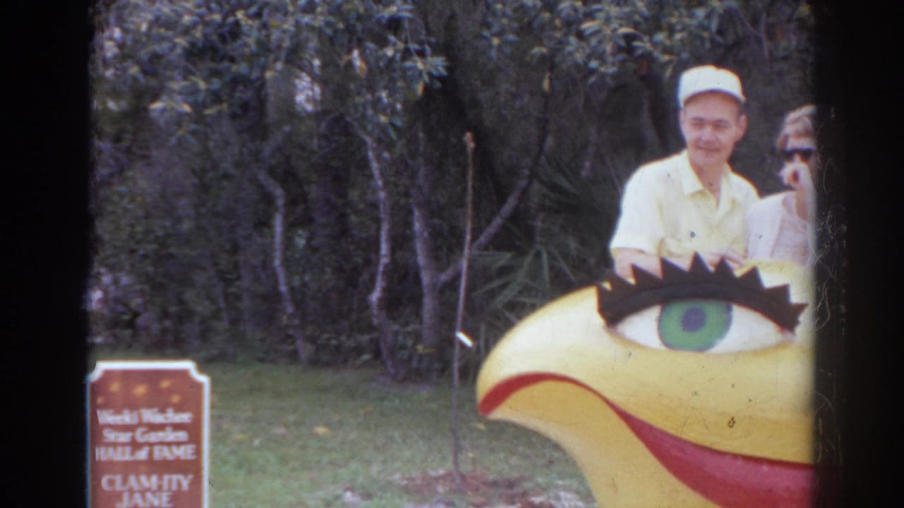In 1966 Norfolk home-movie footage, two adults stand behind a yellow bird-like smiling sculpture at an outdoor attraction.