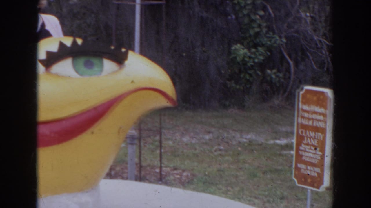 1966 Norfolk home-movie frame showing a large yellow bird figure with a green eye near Orchid Jungle Gardens signage.