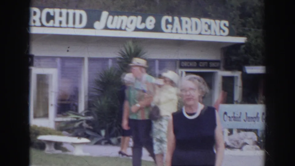 Tourists pass the Orchid Jungle Gardens gift shop in a 1966 Norfolk home movie, with signage, plants, and benches nearby.