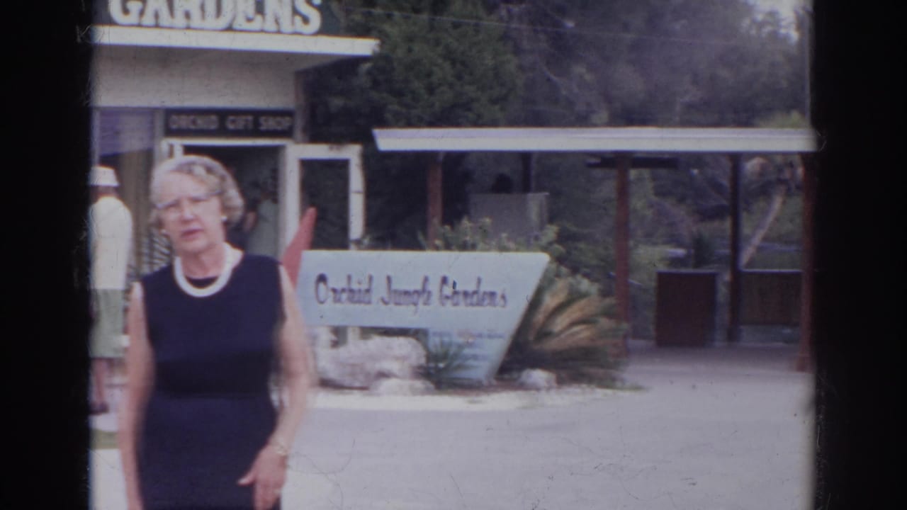 The woman remains near the Orchid Jungle Gardens entrance, with the white garden sign, gift shop, covered walkway, and planting beds visible.