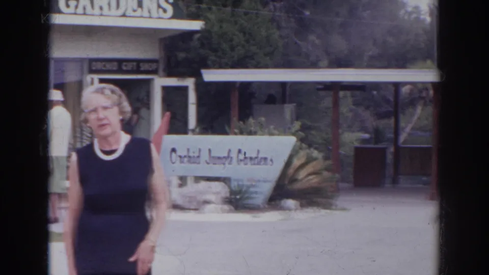 The woman remains near the Orchid Jungle Gardens entrance, with the white garden sign, gift shop, covered walkway, and planting beds visible.