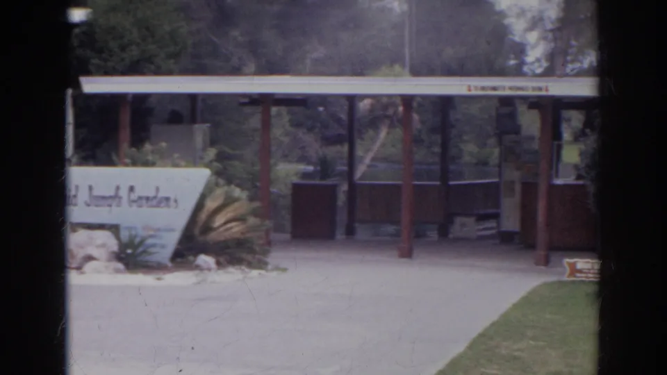 A closer visitor-shot view shows the Orchid Jungle Gardens sign, low planting bed, paved entrance, and covered structure in bright daylight.