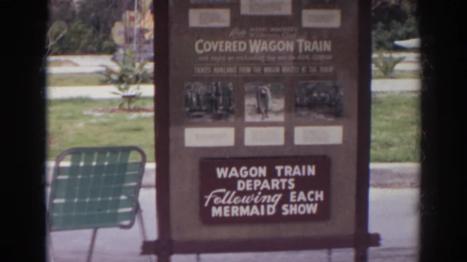 A sign advertising a covered wagon train and departures after each mermaid show stands near a green lawn chair on the garden grounds.