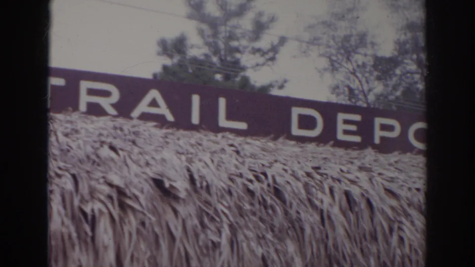 1966 Norfolk home-movie frame showing part of a RAIL DEPOT sign over a thatched roof, with trees and utility lines in the background.