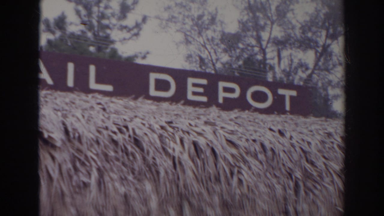 1966 Norfolk frame showing the RAIL DEPOT sign more clearly above a textured thatched roof, with trees visible behind the facade.