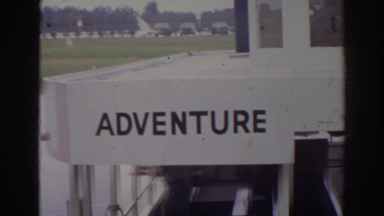 1966 Norfolk frame showing a white ADVENTURE-marked structure, with grass, a roadway, parked cars, and part of the attraction facade visible.