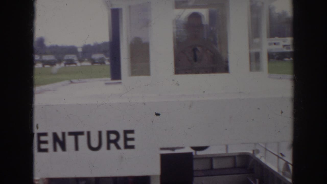 1966 Norfolk frame showing the ADVENTURE structure from closer range, with a person visible through a window and parked cars in the background.
