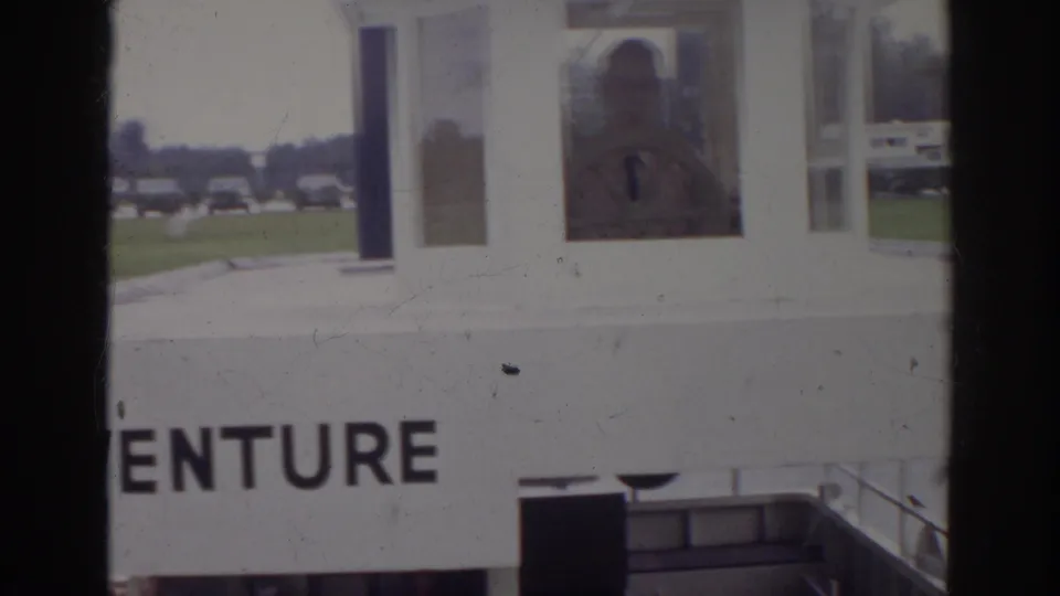 1966 Norfolk frame showing the ADVENTURE structure from closer range, with a person visible through a window and parked cars in the background.