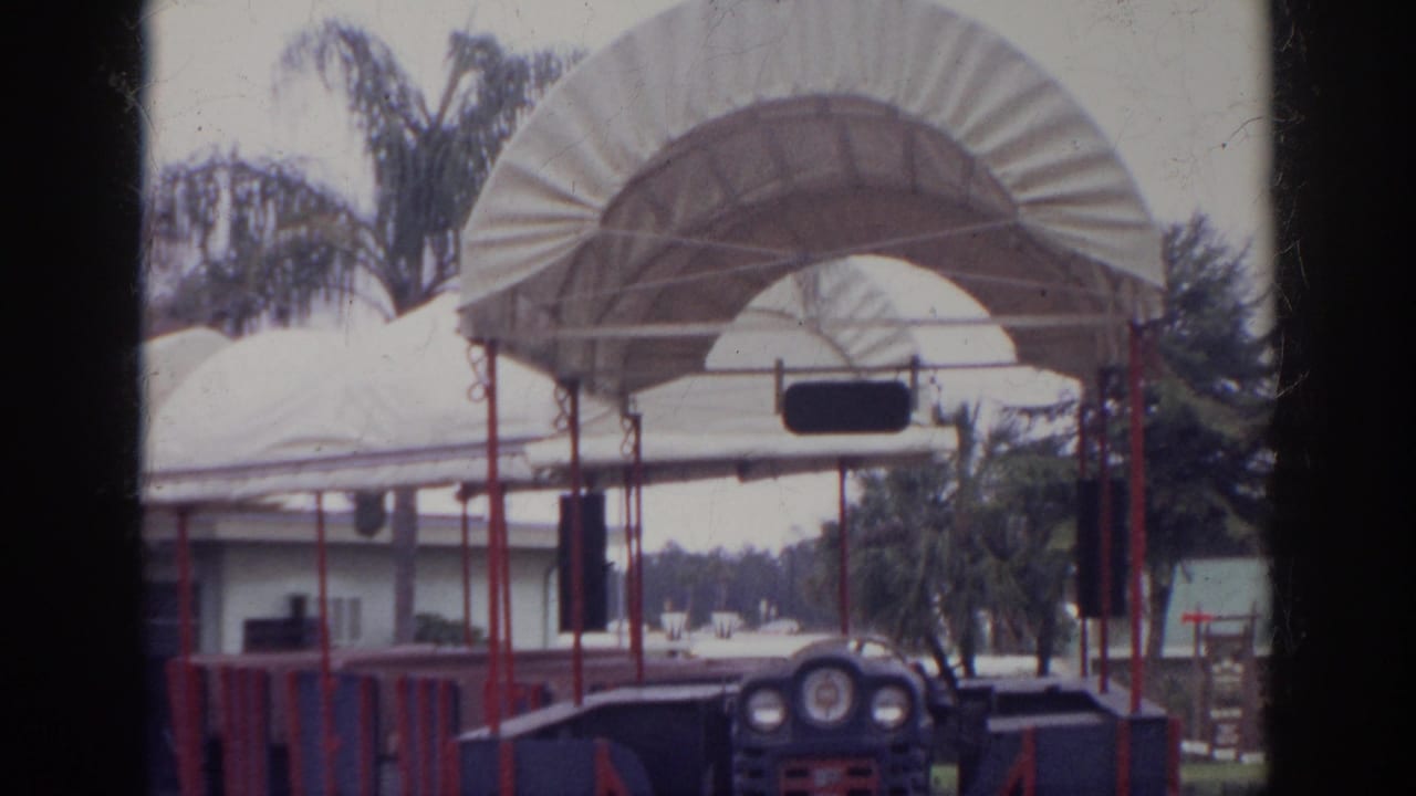 1966 Norfolk home-movie frame showing an empty blue and red tour train near white canopies, palm trees, and a low building.