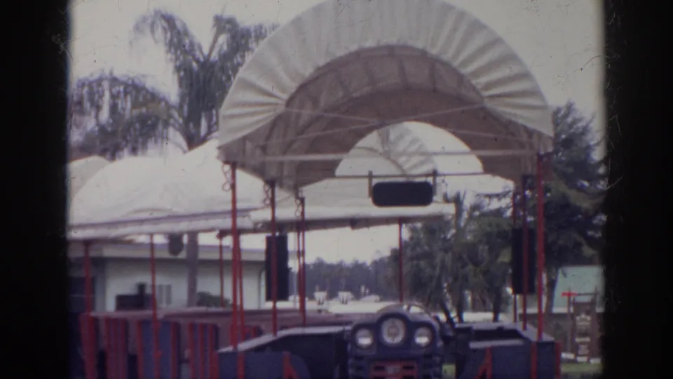 1966 Norfolk home-movie frame showing an empty blue and red tour train near white canopies, palm trees, and a low building.
