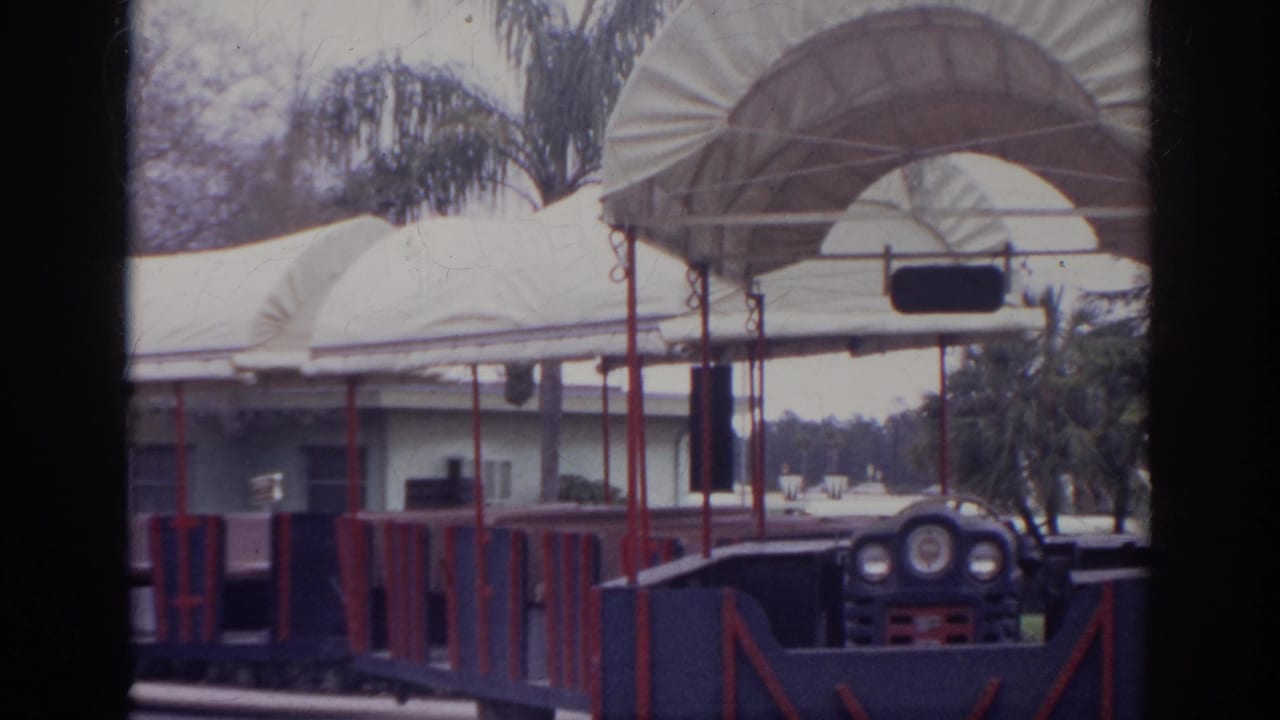 1966 Norfolk frame with the front of an open-air tour train beside white canvas canopies and palm-lined landscaping.