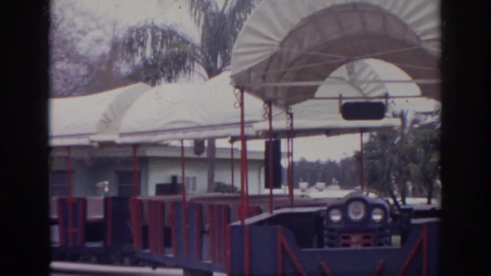 1966 Norfolk frame with the front of an open-air tour train beside white canvas canopies and palm-lined landscaping.