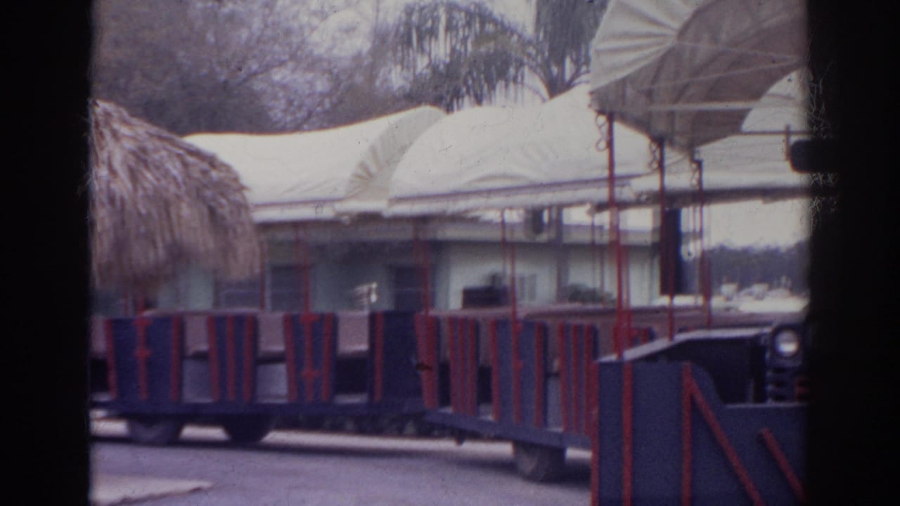 1966 Norfolk home-movie frame showing blue and red open-air cars moving past a low building, canopy roof, and landscaped grounds.