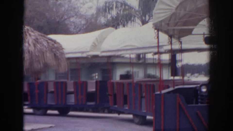 1966 Norfolk home-movie frame showing blue and red open-air cars moving past a low building, canopy roof, and landscaped grounds.