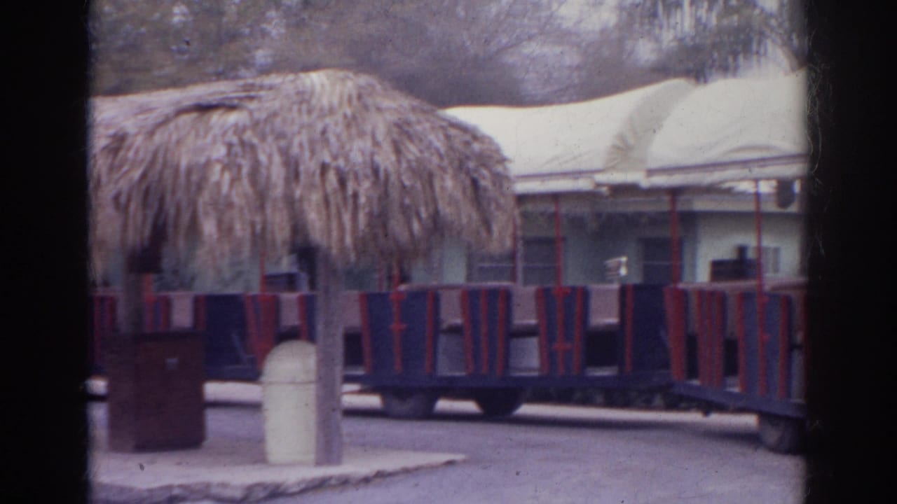 1966 Norfolk frame showing a thatched shelter, empty tour train cars, white canopies, and cloudy park grounds.
