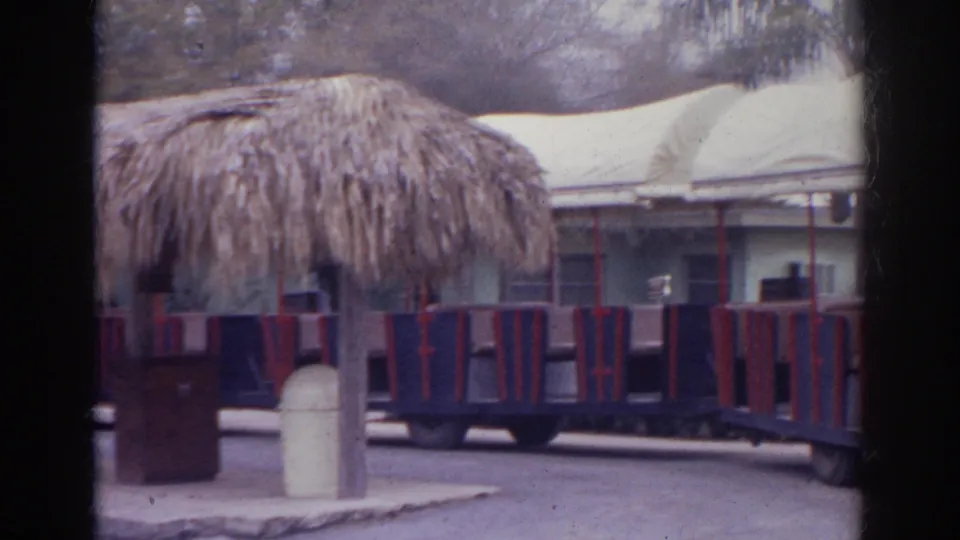1966 Norfolk frame showing a thatched shelter, empty tour train cars, white canopies, and cloudy park grounds.