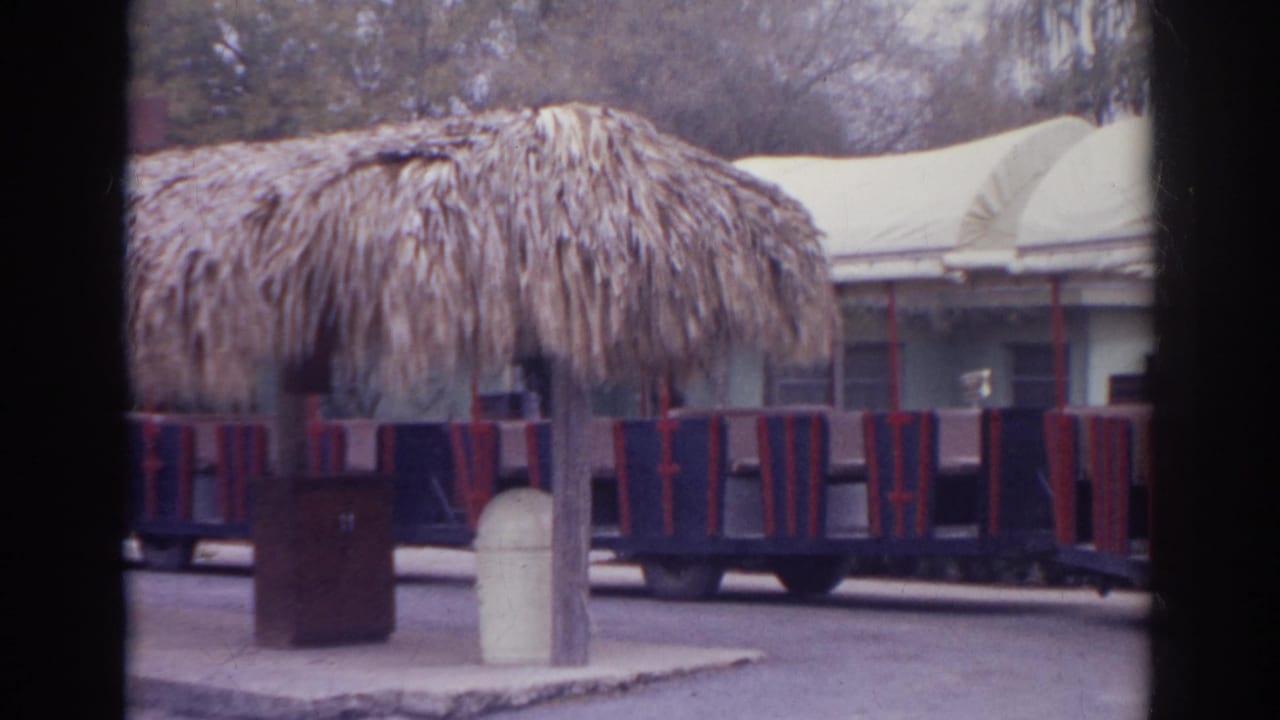 Small open train beside a thatched shelter at a retro park.