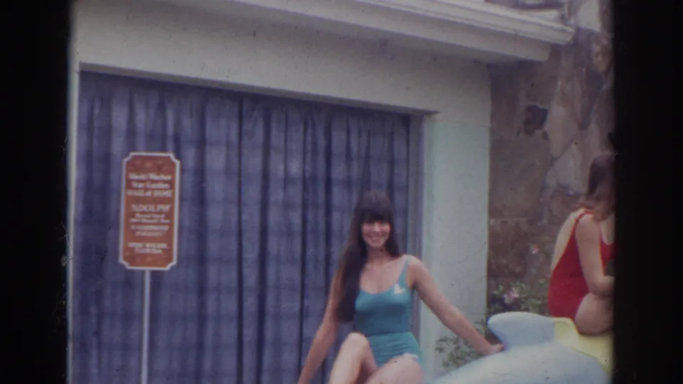 Woman posing on a dolphin statue while another swimsuit-clad visitor is partly visible beside the building.