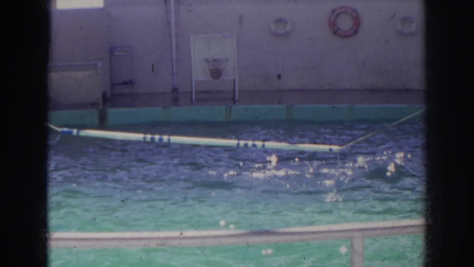 1966 Norfolk home-movie frame showing the pool surface, deck wall, life ring, and a floating or angled show platform.