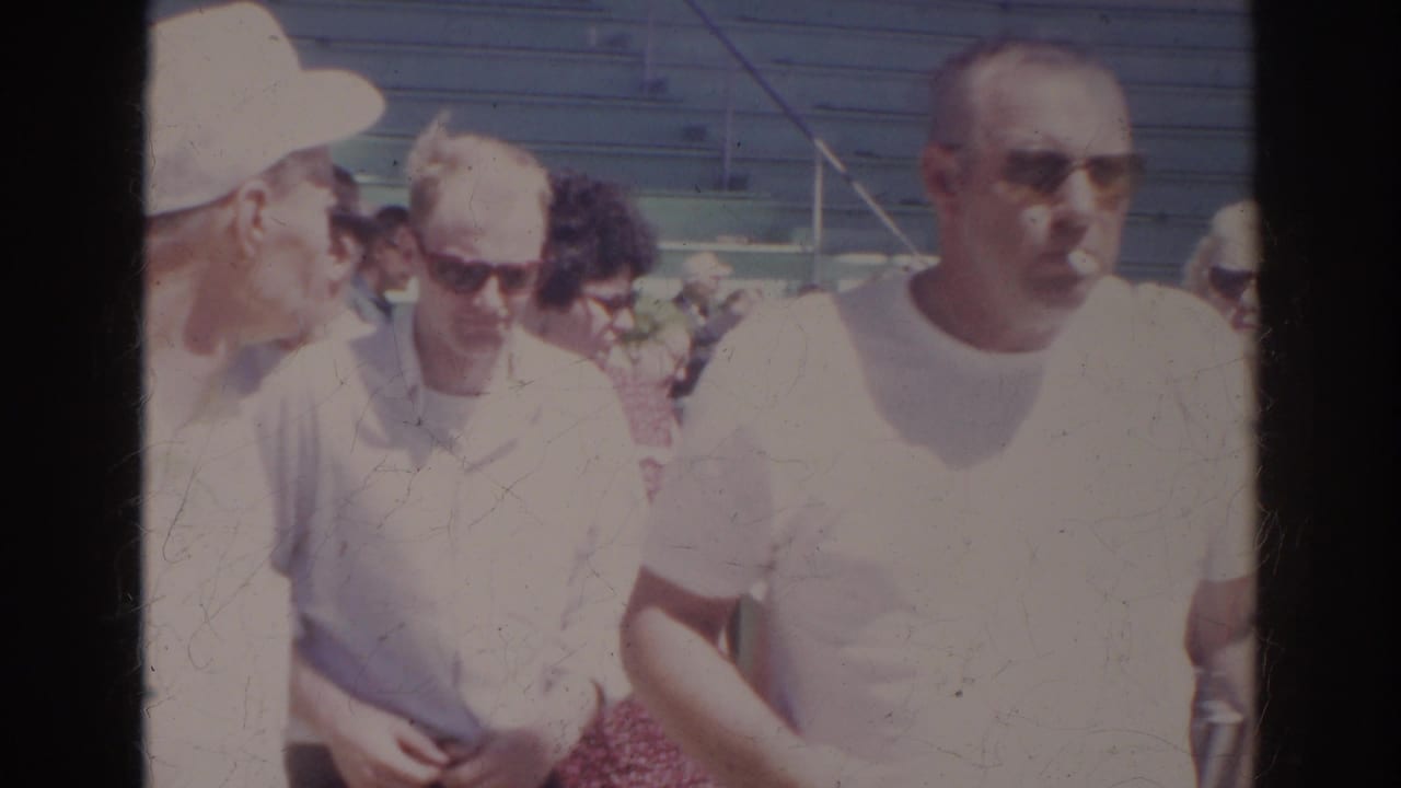 In 1966 Norfolk aquarium home-movie footage, adult visitors in light shirts and sunglasses pass near outdoor seating.