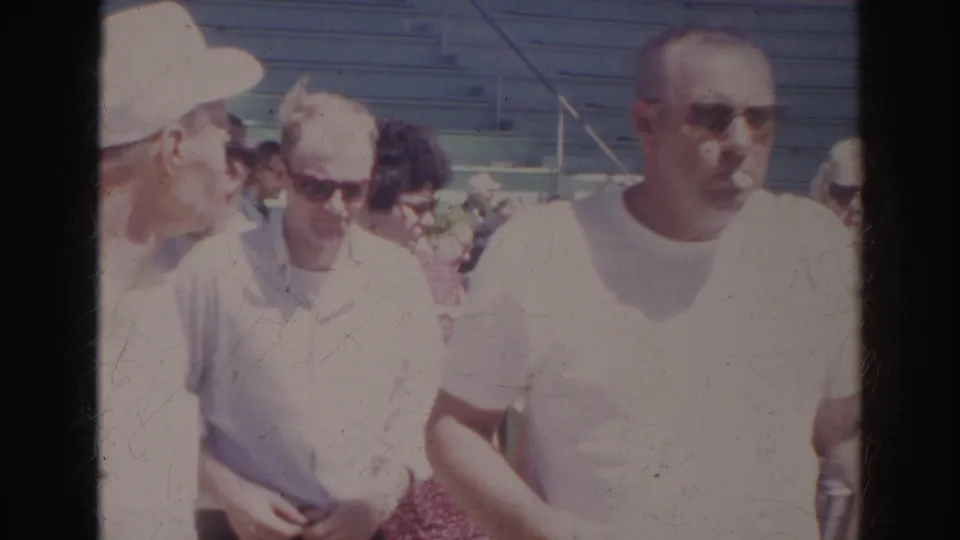 In 1966 Norfolk aquarium home-movie footage, adult visitors in light shirts and sunglasses pass near outdoor seating.