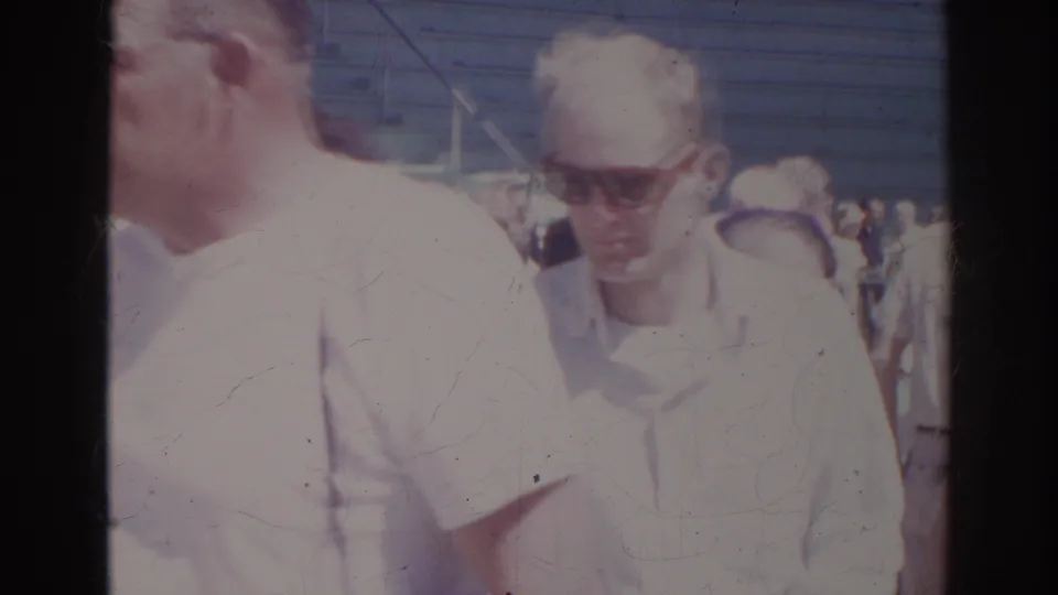 Two adult visitors in light shirts and sunglasses appear in a crowded Norfolk aquarium scene from 1966 home-movie footage.