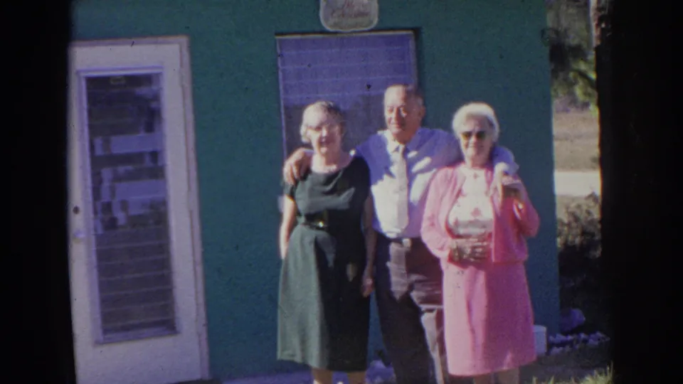 Three older adults face the camera outside a green exterior wall, with a white door and window behind them in Norfolk footage.