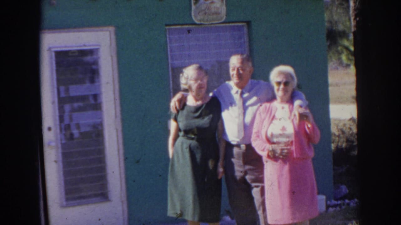 Three adults pose outside a green house near a white door and window.