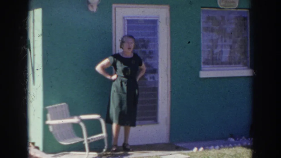 A woman in a dark green dress stands with one hand on her hip outside the green Norfolk house, beside a white glass-panel door.