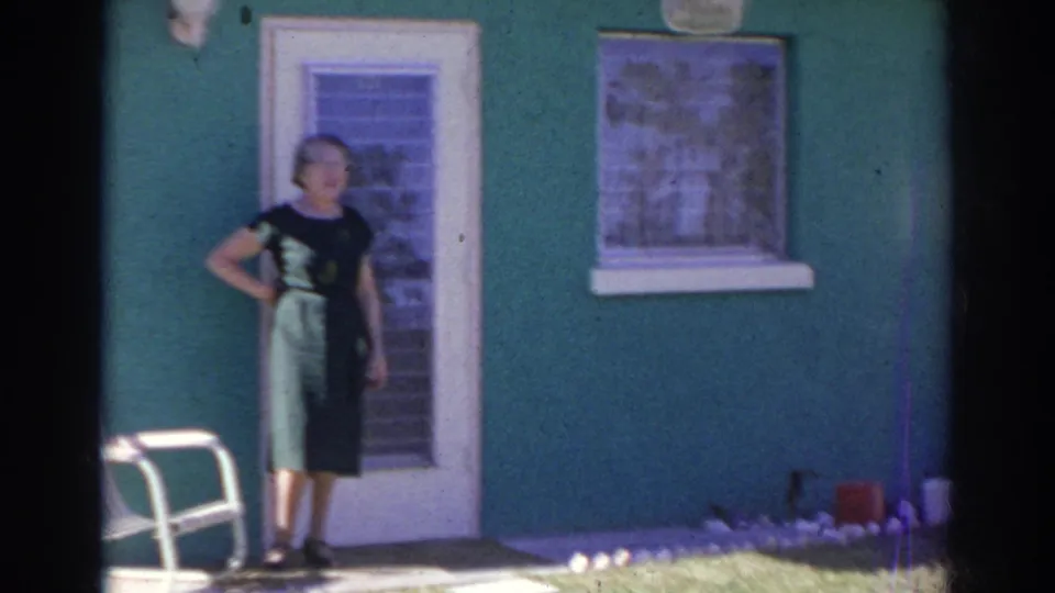 The final frame holds on the woman in the dark green dress outside the bright green house, with a white chair and small garden border nearby.
