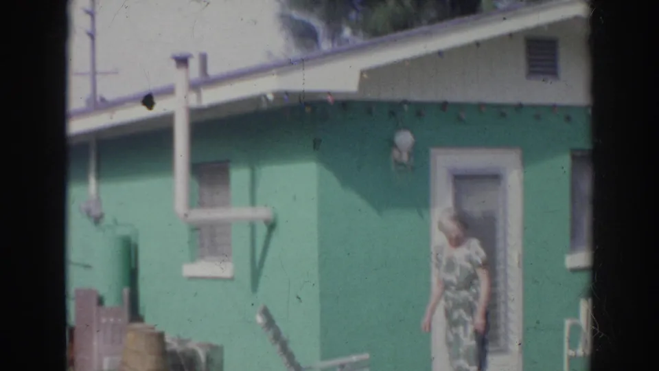 A woman in a patterned dress stands or walks near the door of a green house in the 1966 Norfolk yard scene.