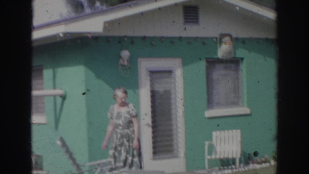 In the Norfolk 1966 home-movie scene, a woman in a patterned dress stands outside a green house near a door, window, and small bench.