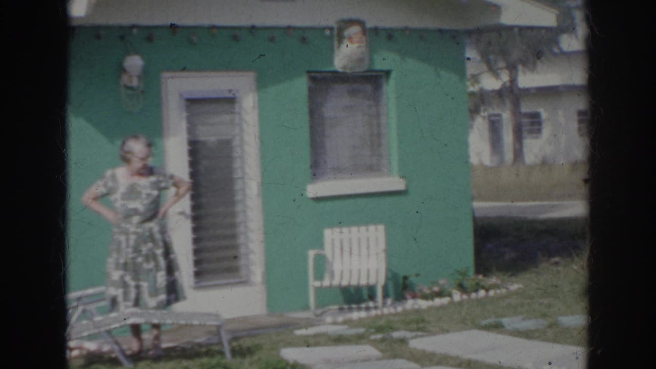 A woman in a patterned dress stands with hands on hips outside the green house, with lawn furniture and stepping stones visible.