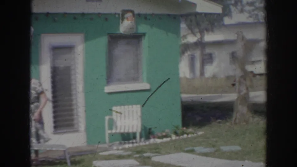 1966 Norfolk home-movie frame showing a teal house, white door, lawn chair, grass, and a woman partly entering from the left edge.
