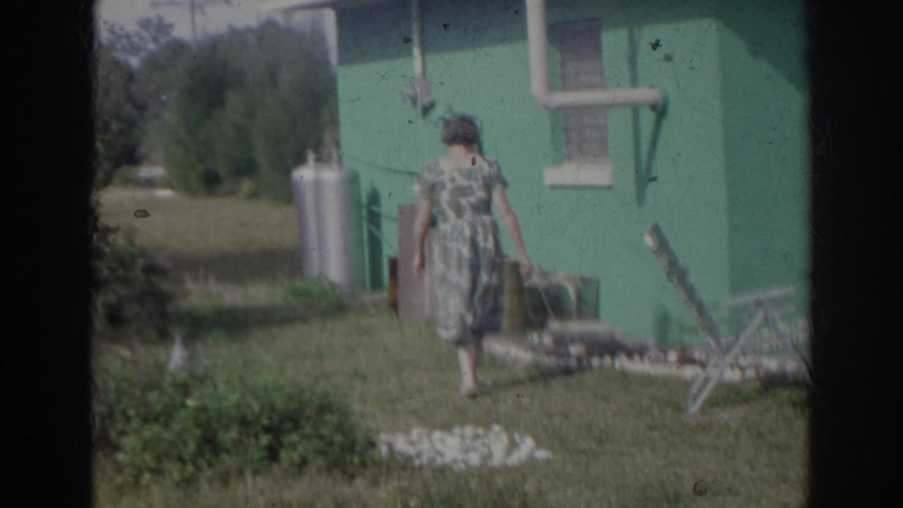 Older woman in a patterned dress walks through a Norfolk yard beside a teal house, with a propane tank and window visible.