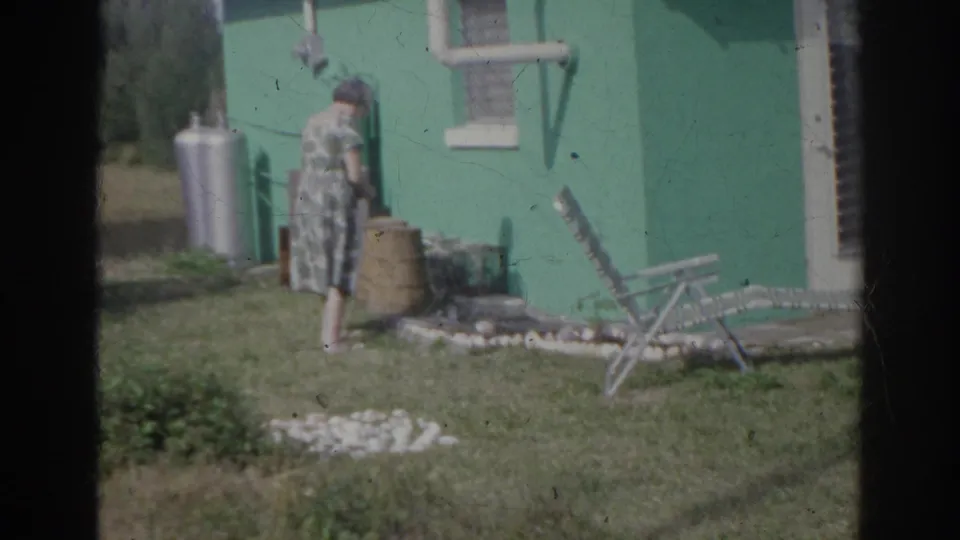 1966 Norfolk frame of an older woman near garden stones beside a teal house, with a propane tank and folding lawn chair in view.