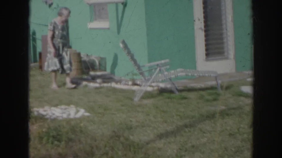 Older woman in a patterned dress moves past lawn chairs and a white door beside the teal house in 1966 Norfolk footage.