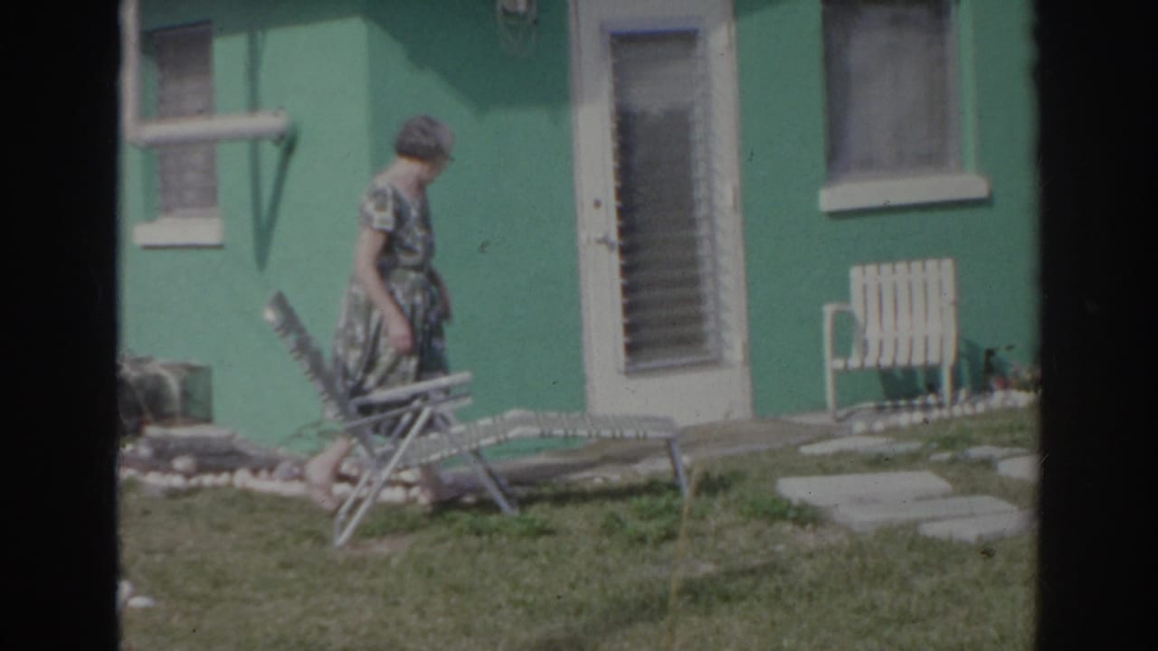 A woman walks across a Norfolk yard in 1966, passing a metal lawn chair outside a bright green house.