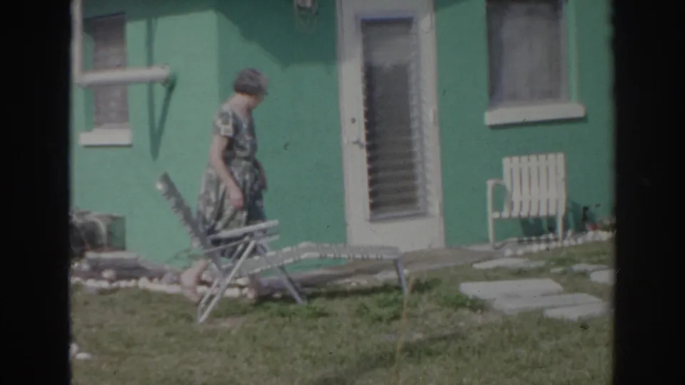 A woman walks across a Norfolk yard in 1966, passing a metal lawn chair outside a bright green house.